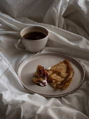 Breakfast in bed, a cup of black tea and thin pancakes with currant jam and sour cream on a beige ceramic plate on a soft white bed in the morning light