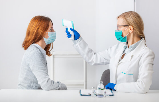 Female medical worker using modern infrared thermometer for checking temperature of caucasian woman. Doctor and patient wearing protective face mask.