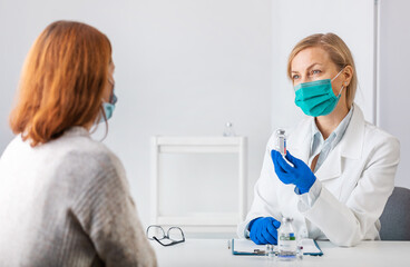 Female doctor in white lab coat, gloves and mask showing to caucasian woman bottle with vaccine while sitting together at medical cabinet. Vaccination of people concept.