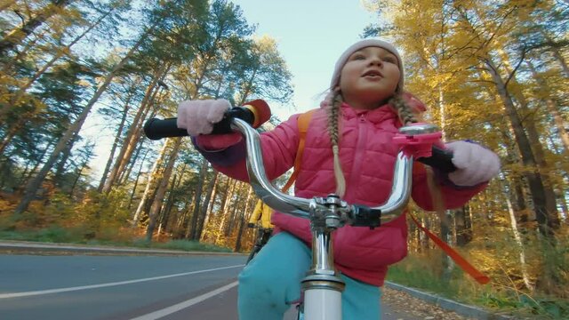 Cheerful Little Girl Rides Bicycle In Autumn Forest, POV Portrait Shot. Cute Caucasian Five-year-old Girl Smiles And Happy, Riding On Bike Path In Park With Family Holding The Handlebars