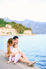 Fototapeta na wymiar Dad, mom and little daughter are sitting on the pier by the sea against the backdrop of the city of Perast and the mountains