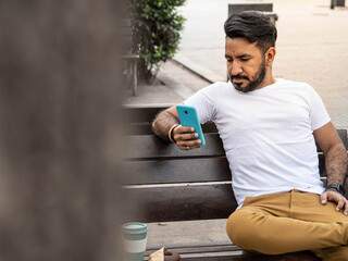latin man sitting on plaza bench looking at cell phone with mug of coffee and donuts, photographed behind tree in foreground