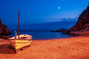 Naklejka premium A boat on a sandy beach in Tossa De Mar Bay lunar night against the backdrop of mountains and blue skies.