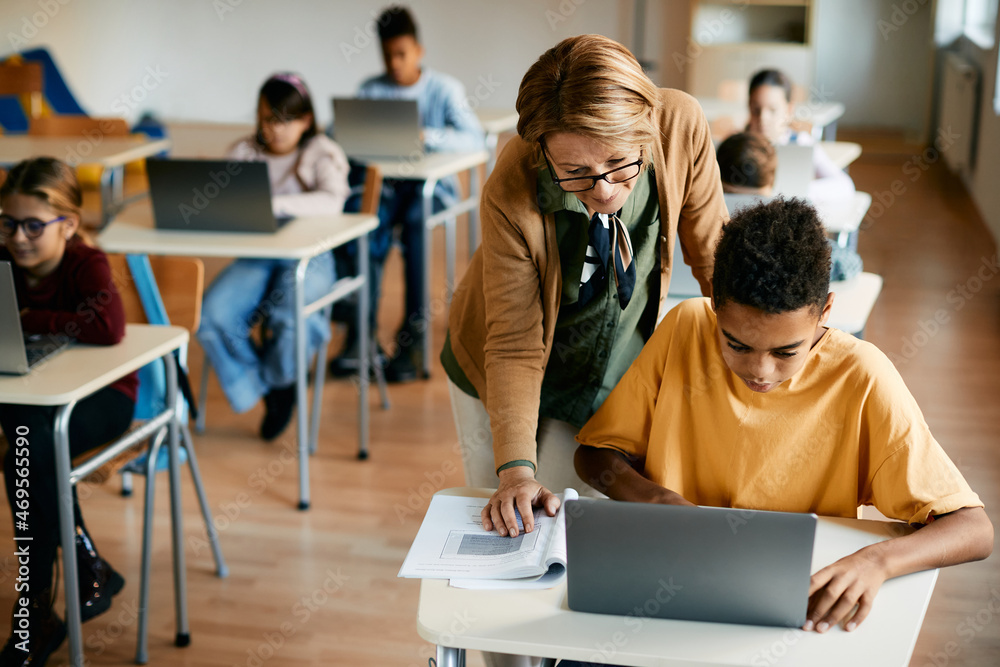 Wall mural African American schoolboy uses laptop with help of his teacher during computer class in classroom.