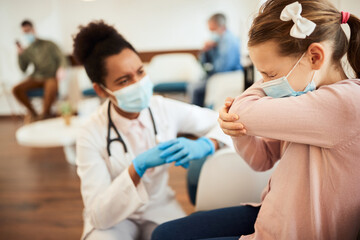 Small girl with face mask sneezes into elbow in waiting room at doctor's office.