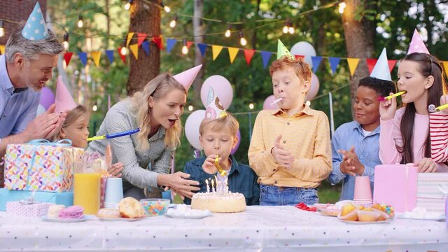 Caucasian cute birthday boy in conus sitting at the table with friends while his mother coming and bringing birthday cake. Holidays concept