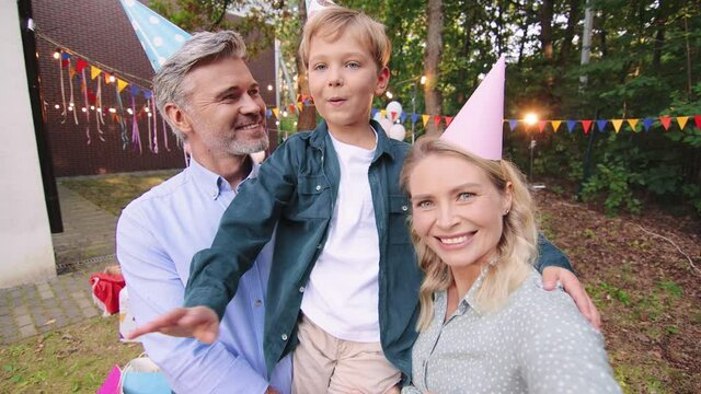 Selfie camera view of the happy family of mother, father and their cute son in party hats standing at the garden and chatting with somebody via video call
