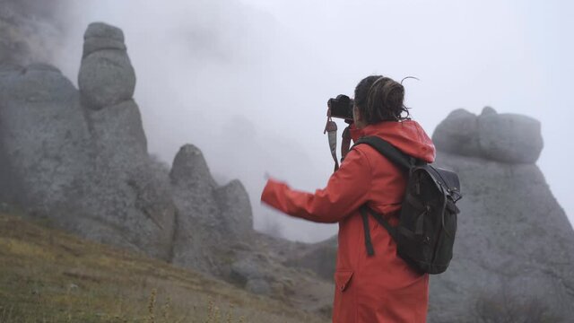 Woman tourist in red raincoat with backpack and photo camera, taking a picture in misty mountains standing between rock cliffs in cold autumn day backside view, tracking shot
