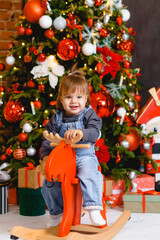 Little beautiful girl child riding wooden toy horse under Christmas tree with gifts