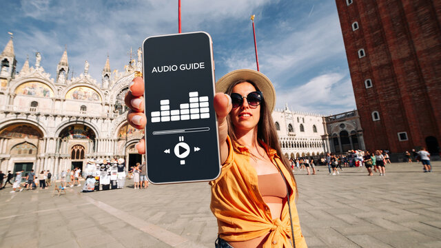Audio tour online app on digital mobile smartphone. Happy young student woman holding phone listening audioguide at San Marco square in Venice, Italy. Simultaneous translation devices.