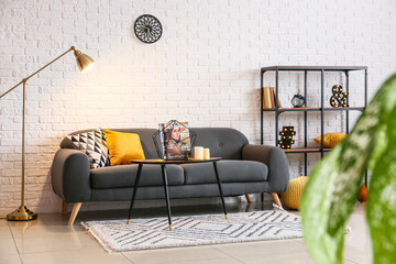 Glowing floor lamp, comfortable sofa, shelf unit, table with candles and magazines in light room