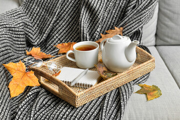 Tray with tea pot, cup of hot beverage and autumn leaves on sofa
