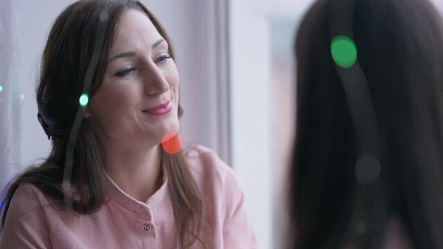 Charming smiling young Caucasian mother talking with adorable little daughter sitting on windowsill waiting for Santa on New Year's eve. Happy woman and girl with Christmas lights at home indoors