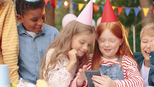 Portrait shot of the cheerful multiracial friends celebrating birthday of the small girl while sitting at the table, watching something funny at the smartphone and laughing