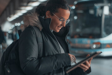 Chica de negocios de cabello oscuro formal con una sonrisa de pie revisando sus apuntes con contratos en la tableta digital en la estación de autobuses por el recinto para tomar apuntes del transporte