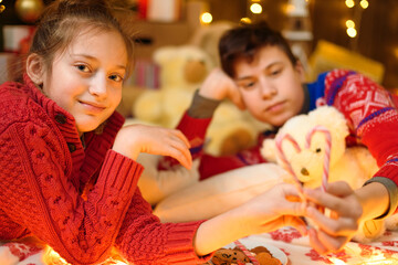 Boy and girl portrait in new year or christmas decoration. They lie and eat sweets and cookies. Holiday lights, gifts and christmas tree decorated with toys.