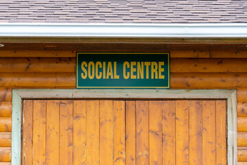A building with orange log siding and a sign with the words social center hung over double wood doors. The sign is green with yellow lettering and references a clubhouse for elderly or aged people.