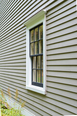 Old tan colored house with white trim and black wood window frame spacers. The glass in the window is wavy and reflects the sun. The exterior wall is made of vintage beige cape cod clapboard siding.