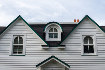 A white wood clapboard siding vintage house with multiple windows. The center window has a curved dormer over the closed glass window in the black shingled portion of the house. The sky is cloudy. 