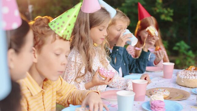 Caucasian pretty birthday girl in conus sitting at the table with friends while eating birthday cake and chatting with each other at the nature
