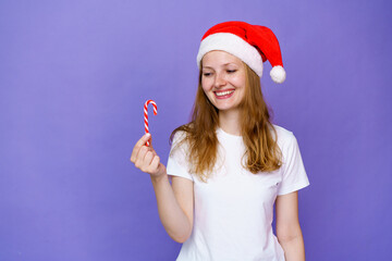 Fototapeta na wymiar A beautiful young girl in white t-shirt and santa hat holds a festive candy in her hand on a lilac background and smiles cutely with happiness. Caucasian woman rejoices at lollipop like a child.