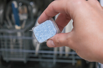 A hand holding blue dishwasher soap tablet in a water-soluble packaging on a dishashind machine background
