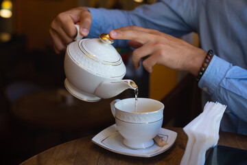 Young businessman in restaurant. Man hands holds white ceramic tea kettle and pouring tea in mug.