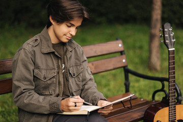Talanted person is talanted in everything. Teenager writes lyrics for a song. Boy using acoustic guitar. Outdoor in autumn time