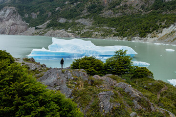 A woman stands on the green hill overlooking a lake with a large iceberg on it, Torres del Paine...