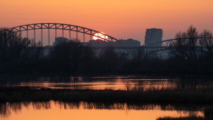 Fototapeta na wymiar Setting sun behind the bridge over the Waal (Rhine) river in Nijmegen against an orange evening sky