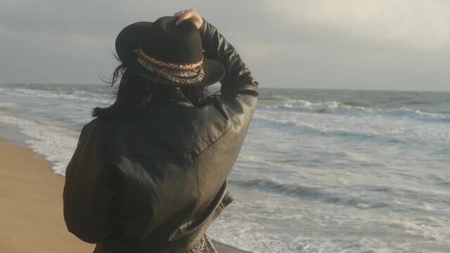 Portrait of romantic brunette woman in hat and leather jacket walking on the beach in autumn at sunset or sunrise 