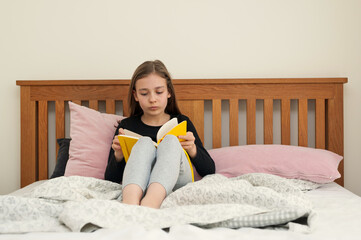 Young school age girl sitting on bed in bedroom and reading from yellow textbook. School homework.