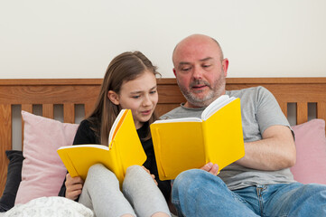 Young school age girl sitting together with her father and reading from yellow textbook. Discussing and solving school homework.