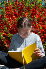 Young school age girl reading from yellow textbook in park against bright red flowers background.