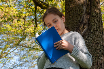 Young school age girl reading from blue textbook in park.