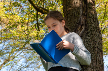 Young school age girl reading from blue textbook in park.