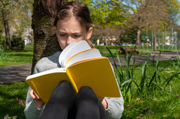 Young school age girl reading from yellow textbook in park.