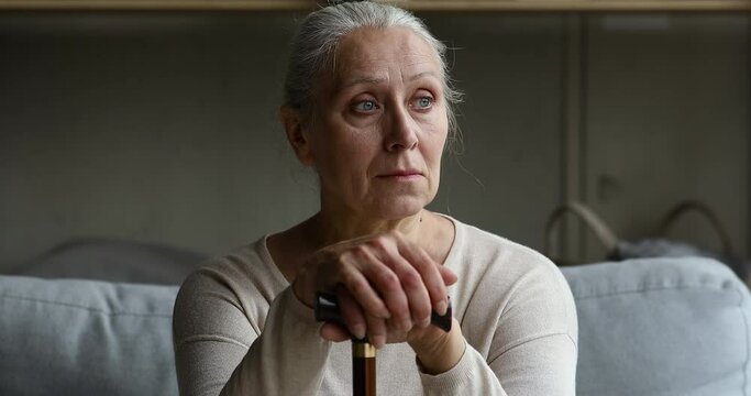 Sad old lady having disability sit on couch alone lean on walking cane look aside think of health problem. Sick lonely mature female feel abandoned unhappy ponder on difficulties of retirement life