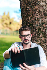 Fototapeta premium Young intelligent man wearing eyeglasses is reading book leaning on a tree in the city park