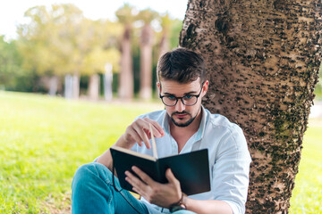 Young intelligent man wearing eyeglasses is reading book leaning on a tree in the city park
