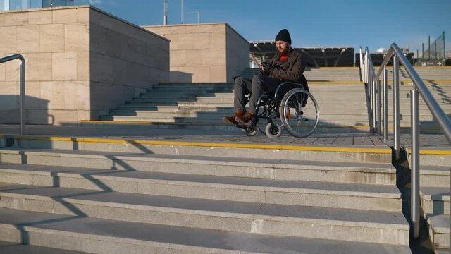 man with paralysis of bottom body part is sitting in wheelchair in city, messaging by smartphone