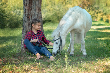 a teenage girl walks with a white pony in the forest, they sit by a tree while the horse grazes