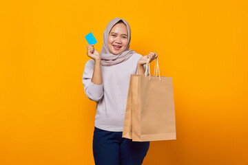 Portrait of cheerful asian woman holding shopping bag and showing credit card over yellow background