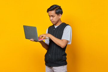 Portrait of handsome young man using laptop looking serious doing office work on yellow background