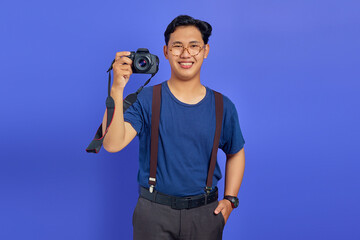 Handsome photographer showing professional camera and smiling cheerfully on purple background