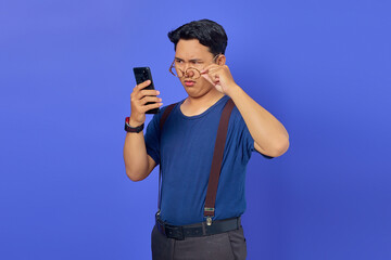 Shocked Asian young man looking at smartphone screen and taking off glasses on purple background