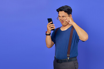 Shocked Asian young man looking at smartphone screen and taking off glasses on purple background