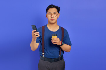 Sad handsome young man holding smartphone and cup of coffee on purple background