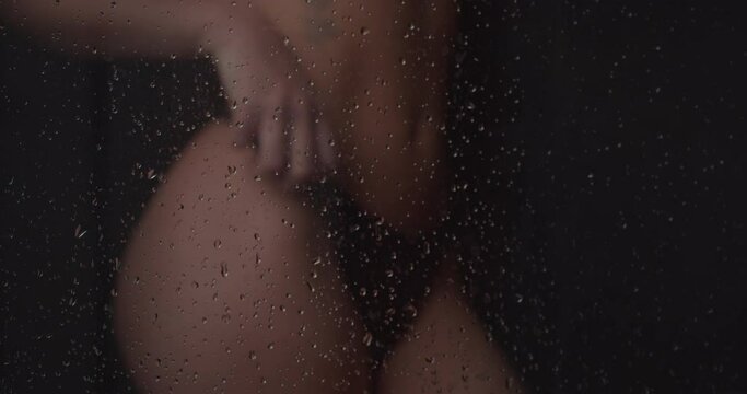 4K close up of an athletic female model in underwear with flat belly taking a shower behind the shower screen while the shower drops fall on her body. Concept of sexy women wet in the shower.