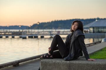 Biracial teen girl sitting by lake at sunset, wearing jacket